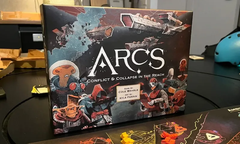 Die Arcs-Schachtel auf einem Spieltisch, davor das Ambitionen-Tableau und bunte Spielfiguren