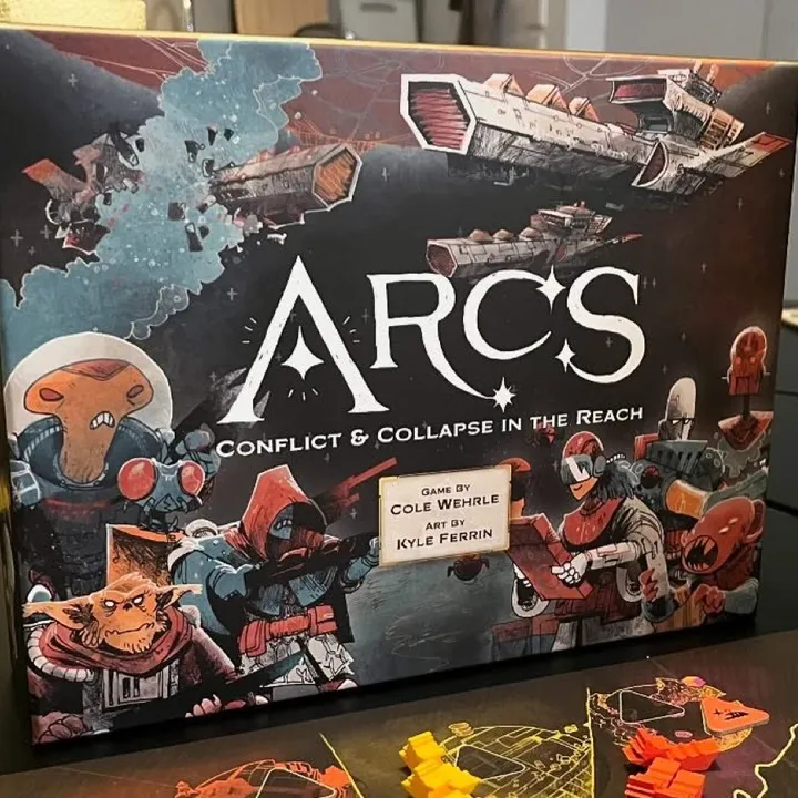 Die Arcs-Schachtel auf einem Spieltisch, davor das Ambitionen-Tableau und bunte Spielfiguren