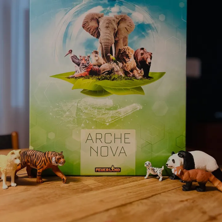 Die Schachtel von Arche Nova auf einem Holztisch, umringt von Spielzeugtieren: ein Tiger, ein Panda, ein Roter Panda und ein Dalmatiner