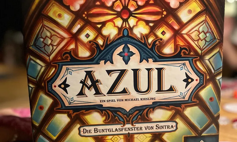 Die Schachtel von Azul: Die Buntglasfenster von Sintra (deutsche Ausgabe) auf einem Holztisch, im Vordergrund orangefarbene Glasteile