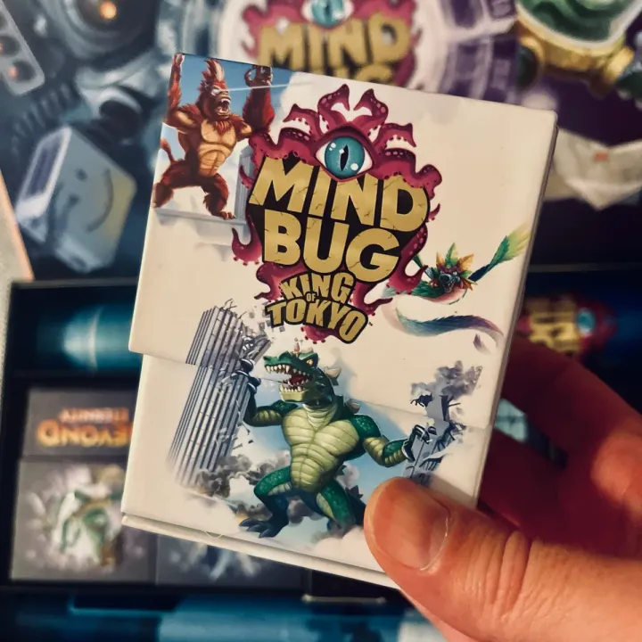 Die Box Mindbug x King of Tokyo in der Hand, dahinter die Mindbug-Sammelbox