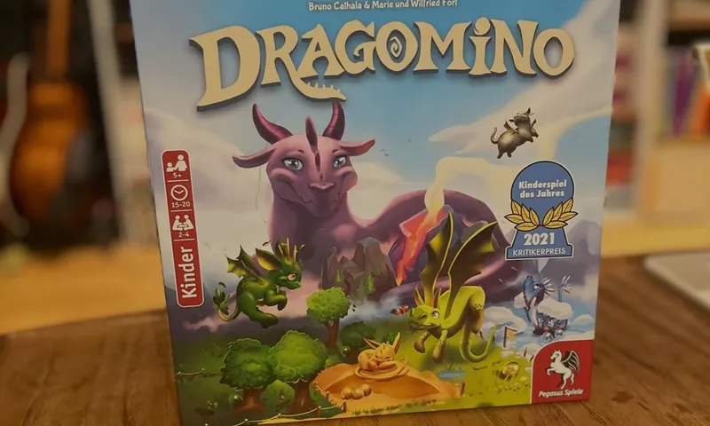 Die deutsche Dragomino-Schachtel mit einem lila Drachen auf dem Cover und der Auszeichnung Kinderspiel des Jahres 2021, davor Plättchen und Dracheneier