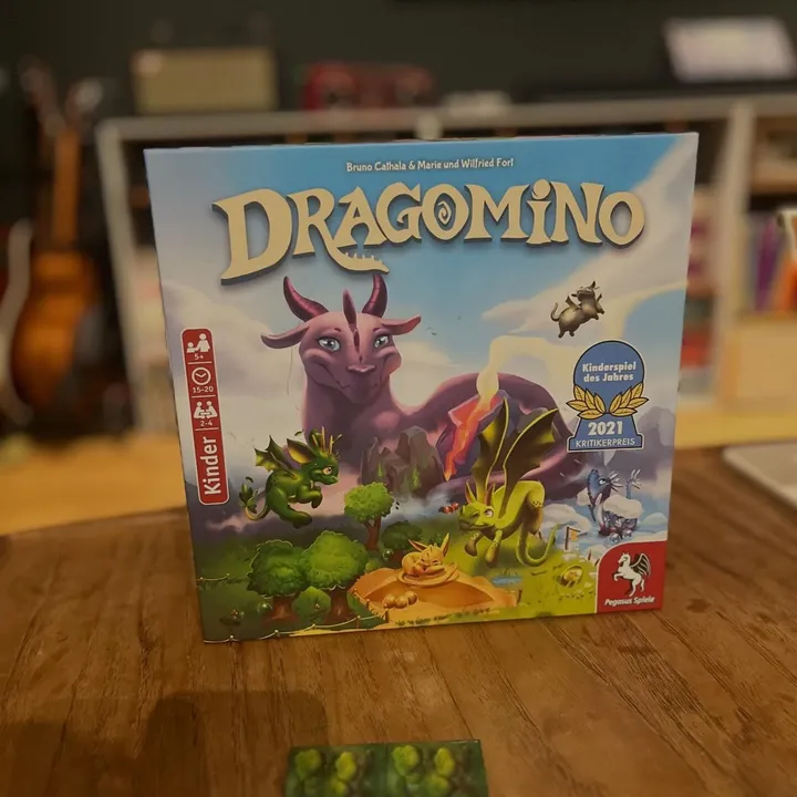 Die deutsche Dragomino-Schachtel mit einem lila Drachen auf dem Cover und der Auszeichnung Kinderspiel des Jahres 2021, davor Plättchen und Dracheneier