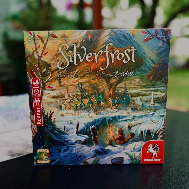 Die Schachtel von Everdell Silverfrost auf einem dunklen Tisch im Freien, Bäume im Hintergrund