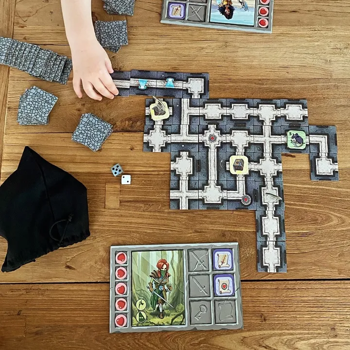 Eine Partie Karak von oben: Dungeon-Plättchen auf einem Holztisch, Heldentafeln, Würfel und eine Kinderhand, die ein Plättchen anlegt