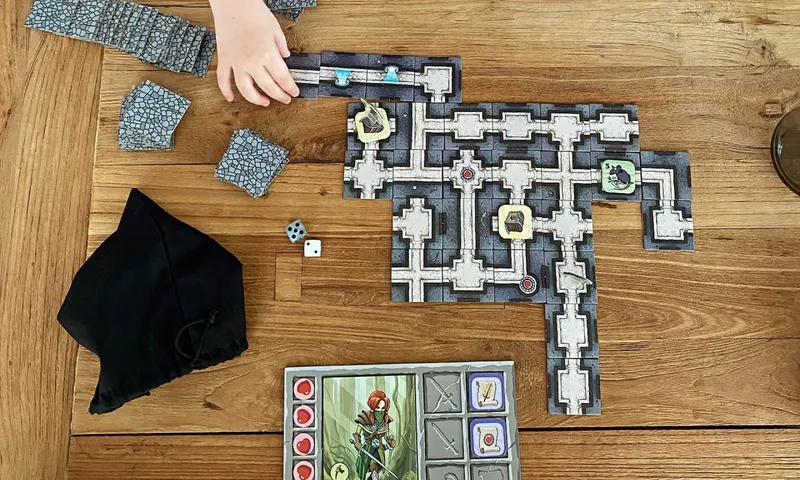 Eine Partie Karak von oben: Dungeon-Plättchen auf einem Holztisch, Heldentafeln, Würfel und eine Kinderhand, die ein Plättchen anlegt