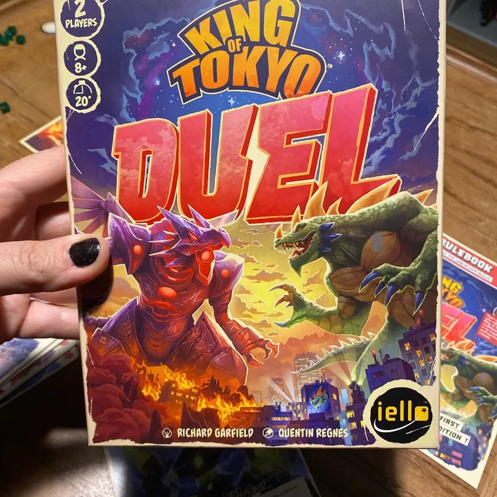 A hand holding the King of Tokyo: Duel box above a game table, with the rulebook and cards in the background