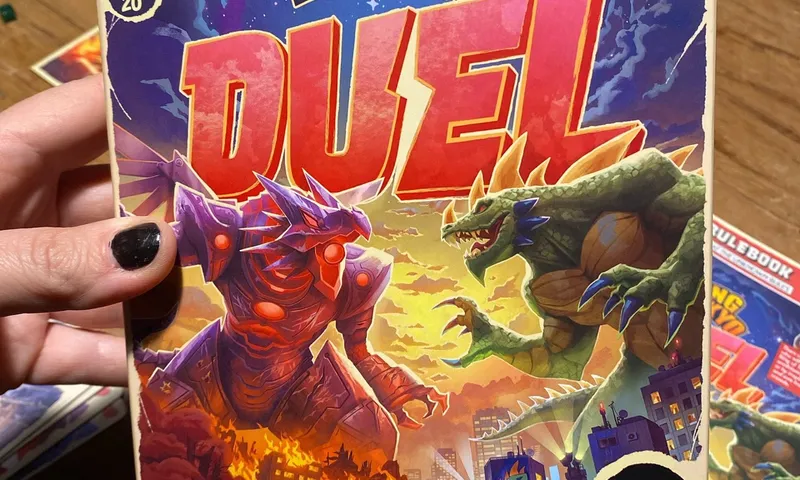 Eine Hand hält die Schachtel von King of Tokyo: Duel über einen Spieltisch, im Hintergrund Regelheft und Karten