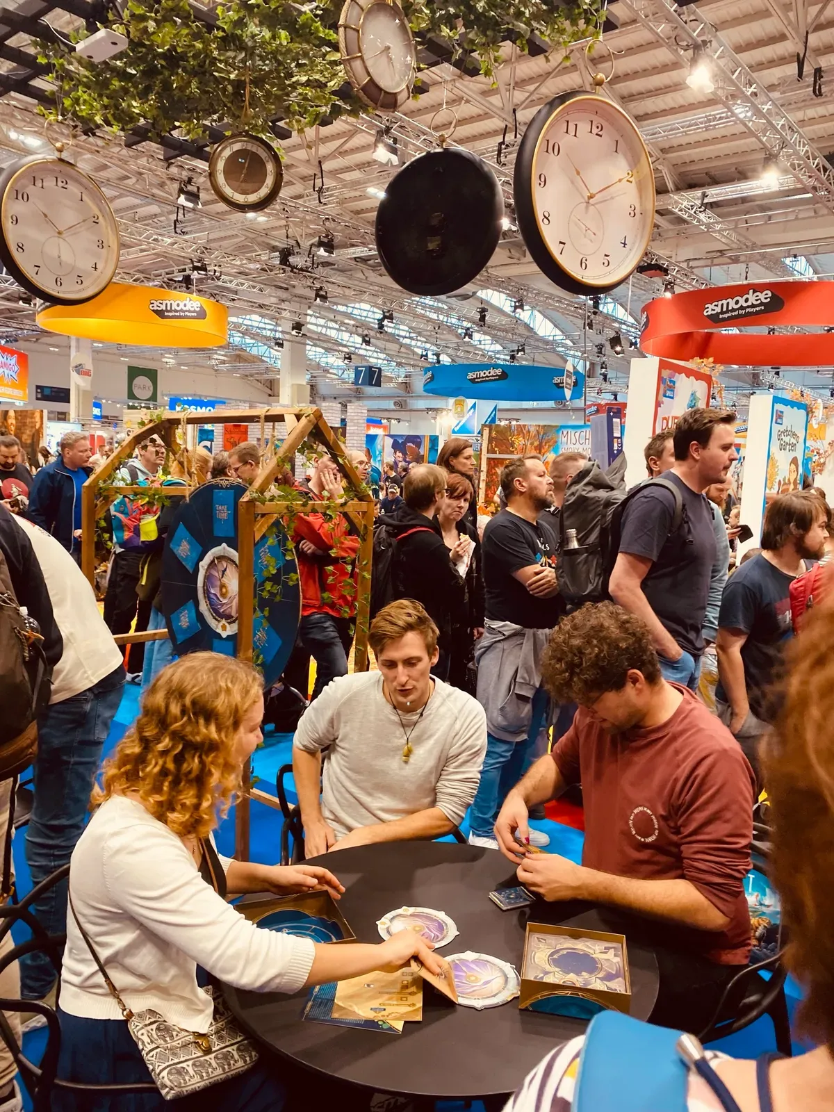 Ein Spieltisch im Asmodee-Bereich, über den Köpfen hängen Uhren als Deko