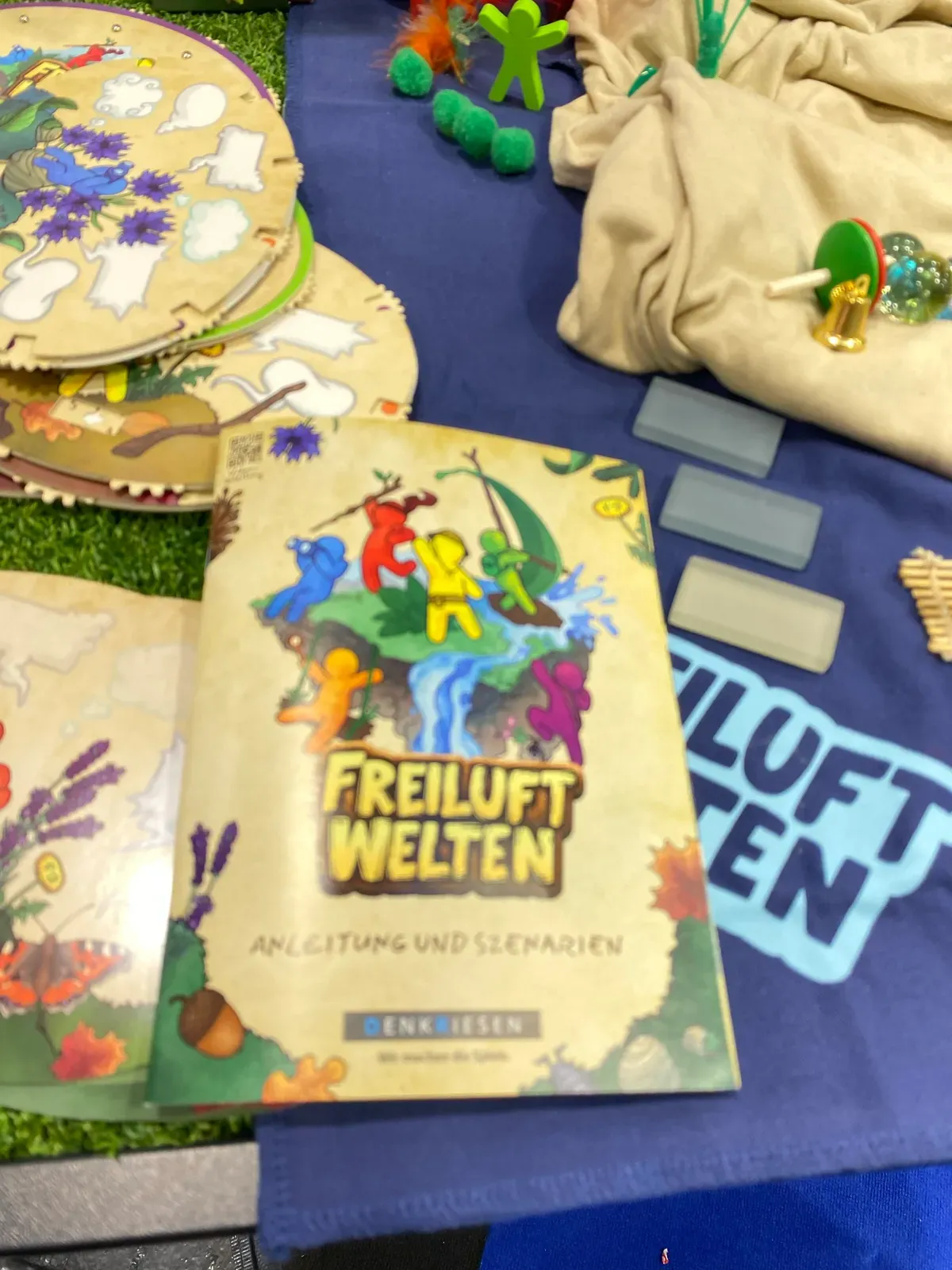 Das Anleitungsheft von Freiluftwelten auf einem Stand mit Spielfiguren und Stoffen