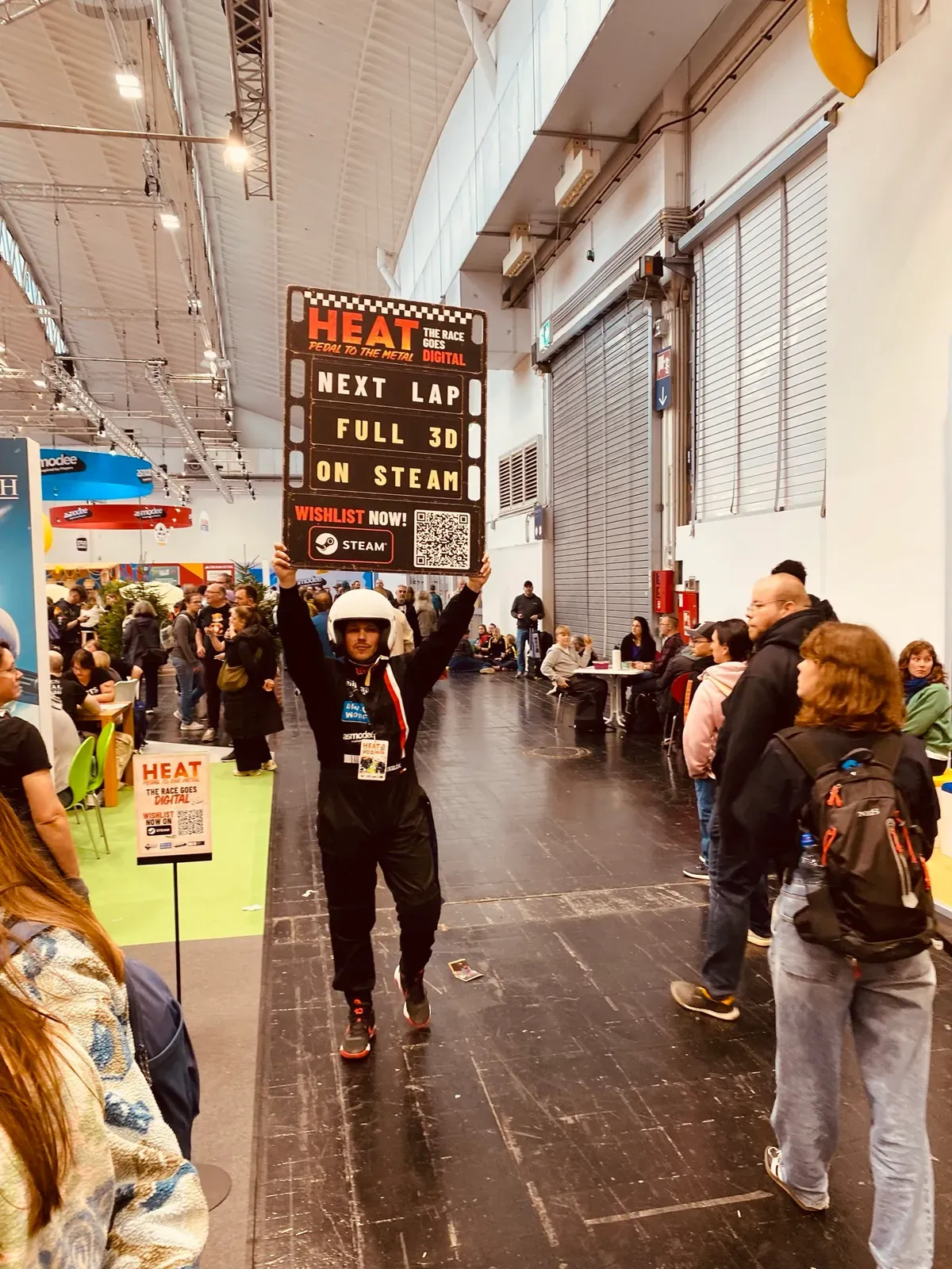 Ein Darsteller im Rennanzug mit Schild „Heat: Next Lap, full 3D on Steam“ im Gang