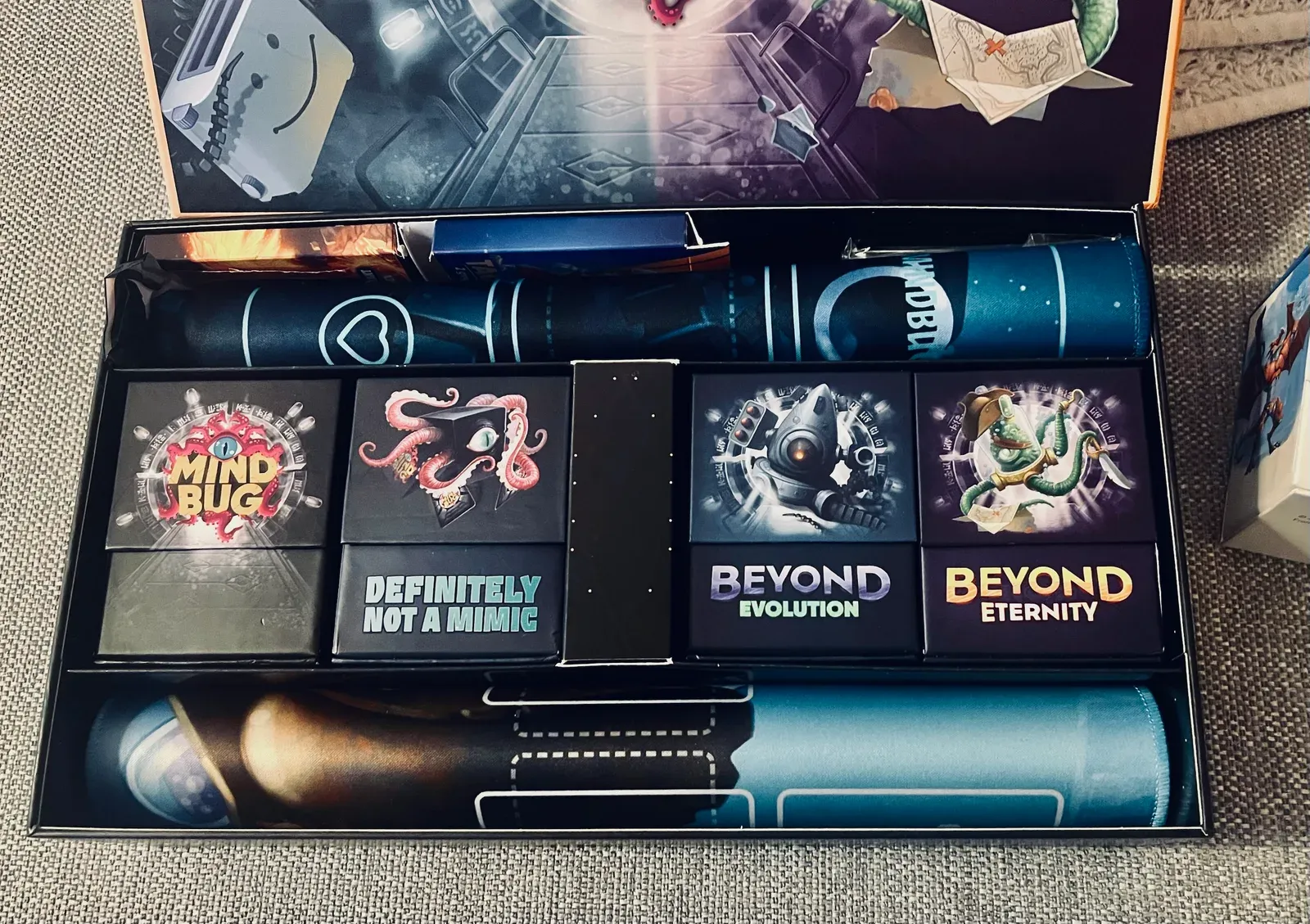 Blick in unsere Mindbug-Sammelbox: die Boxen Mindbug, Beyond Evolution und Beyond Eternity, dazu die Box Definitely Not a Mimic mit unseren Erweiterungen, zwischen zwei gerollten Spielmatten