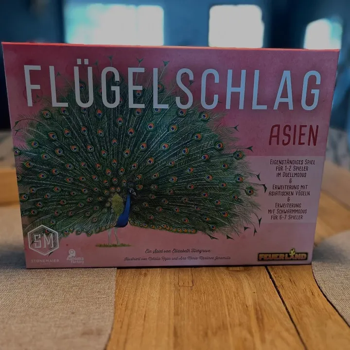 Die deutsche Ausgabe von Wingspan Asia (Flügelschlag Asien) mit einem Pfau auf der Schachtel, auf einem Tisch stehend